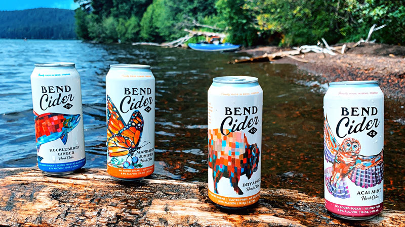 4 canned flavors of Bend Cider on a log by the river
