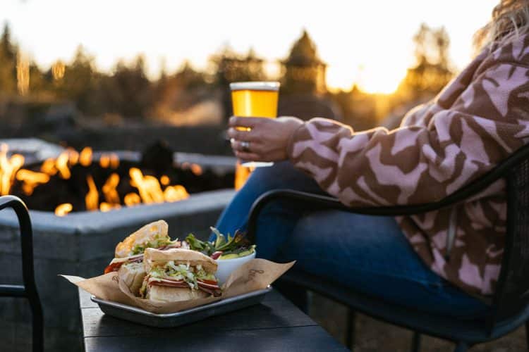 Person holding beer by fireplace with sandwich on plate