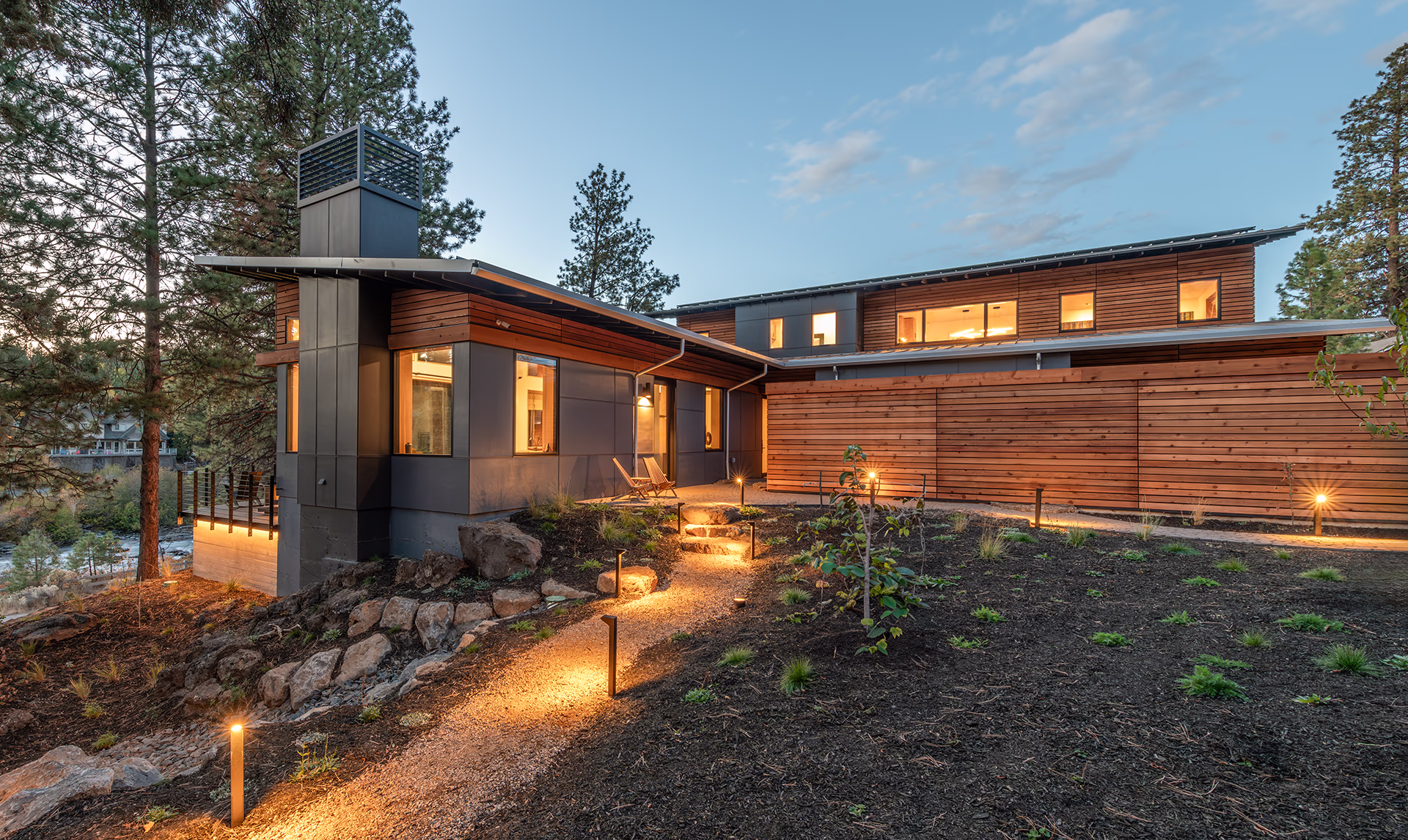 Modern wood and metal Bend home glowing at dusk beside a landscaped path and pine trees.